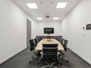 Een vergaderruimte met een lange tafel, zwarte stoelen, een flatscreen televisie en een plafondlamp.