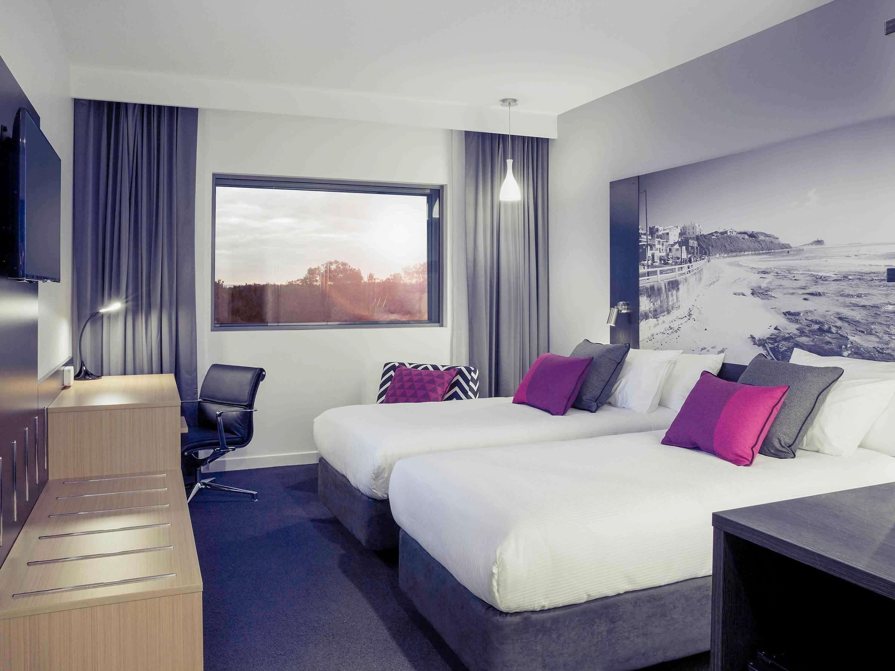 A modern hotel room with two beds, a desk, a chair, a TV, and a large window with a beach view.