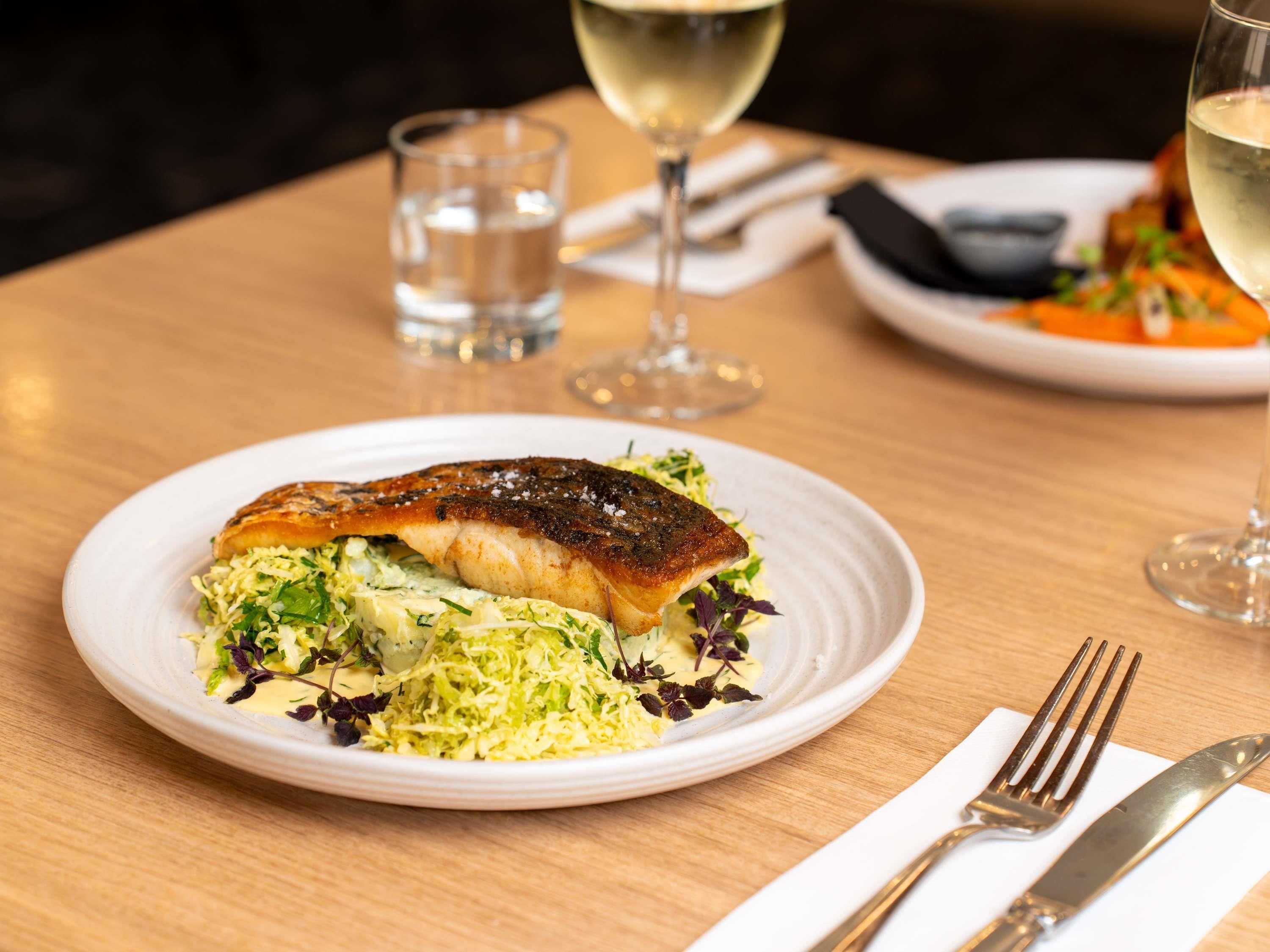 A plate of food with a fish fillet, greens, and a glass of white wine on a wooden table.