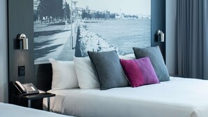 Une chambre d’hôtel avec un lit, des oreillers, une table de chevet avec un téléphone et une grande fenêtre donnant sur le front de mer et le paysage urbain.