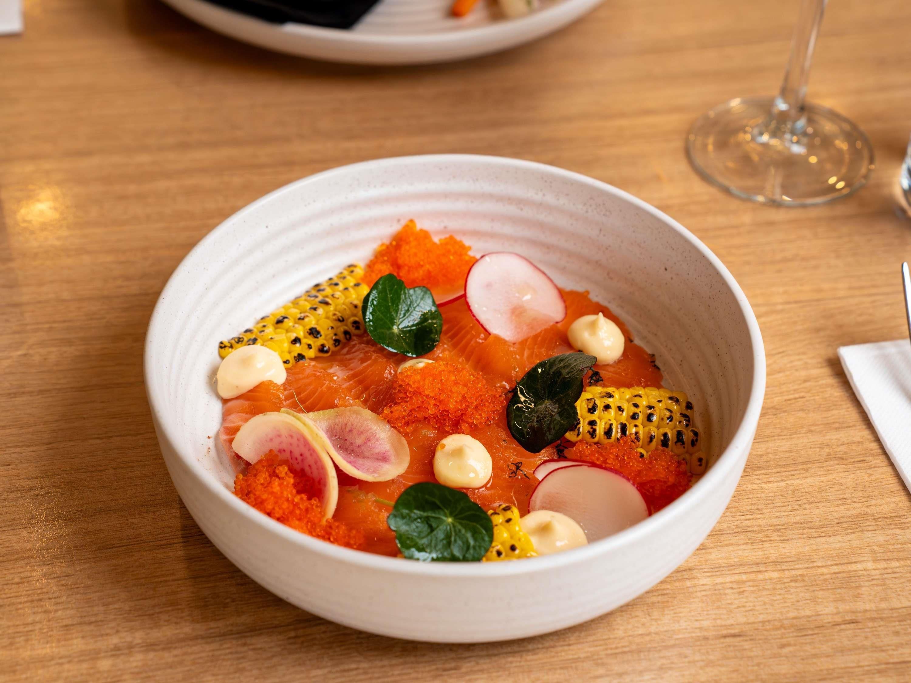A bowl of food with salmon, caviar, corn, and radish on a wooden table.