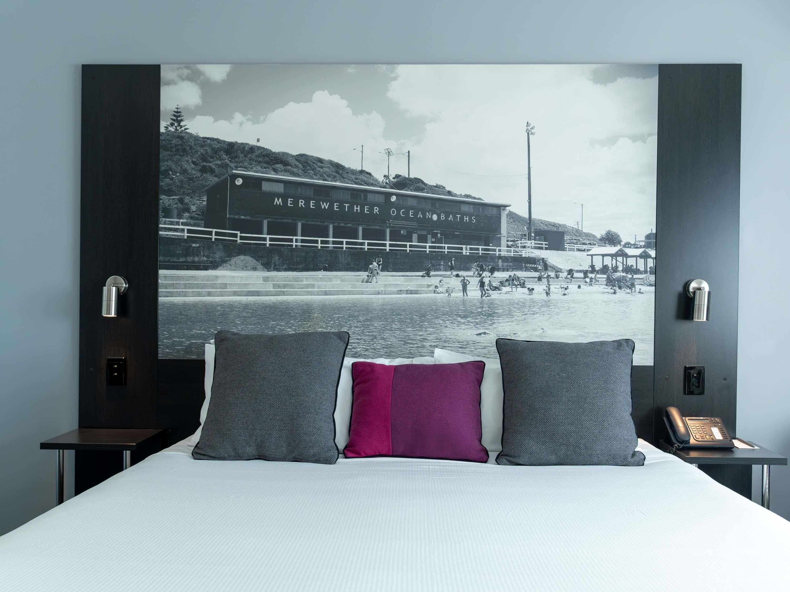 A bedroom with a large bed, two pillows, a nightstand with a phone, and a wall-mounted light.
