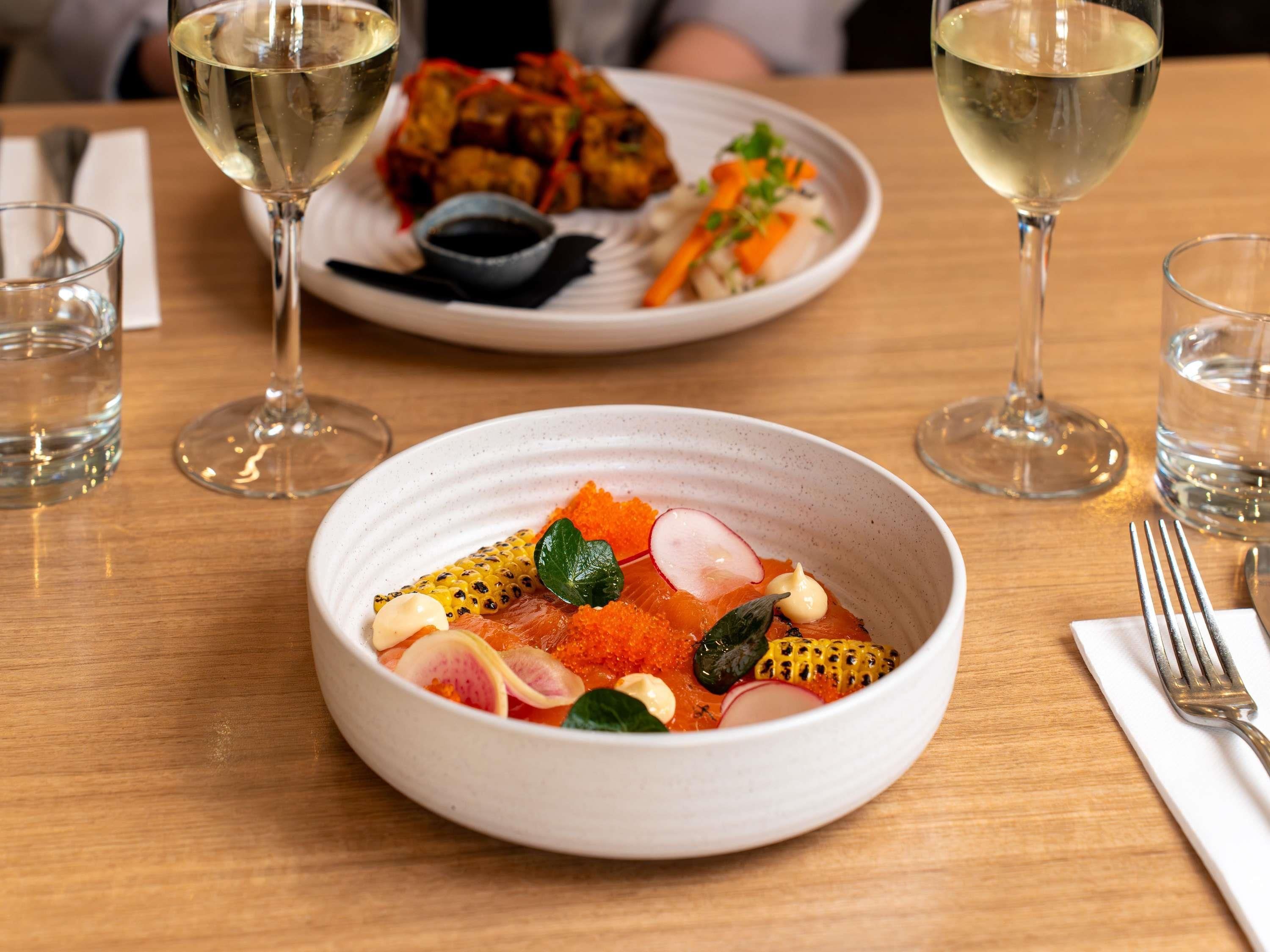 A dining table with a bowl of food, two glasses of white wine, and a fork on a napkin.