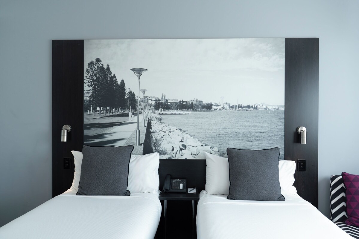 Habitación de hotel con cama, dos almohadas, mesita de noche con teléfono y una amplia vista en blanco y negro de un paisaje urbano.