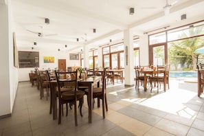 A spacious indoor dining area with multiple wooden tables and chairs, large windows, and ceiling fans.