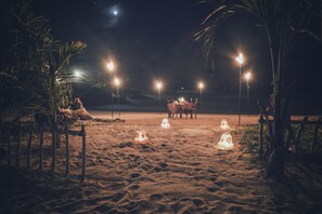 Une plage avec une table et des chaises placées sous quatre lampes, entourées de palmiers et de sable.