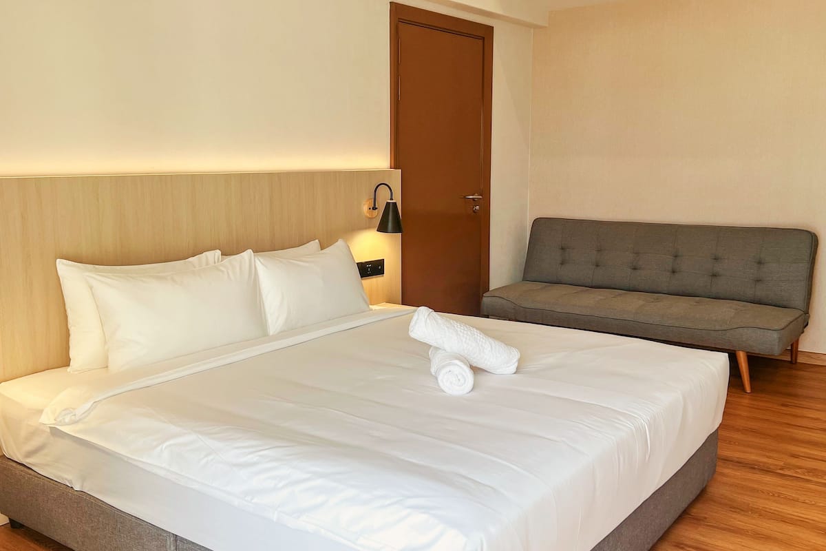 A neatly made bed with white linens and a rolled towel, a grey upholstered sofa, and a wooden door in a hotel room.