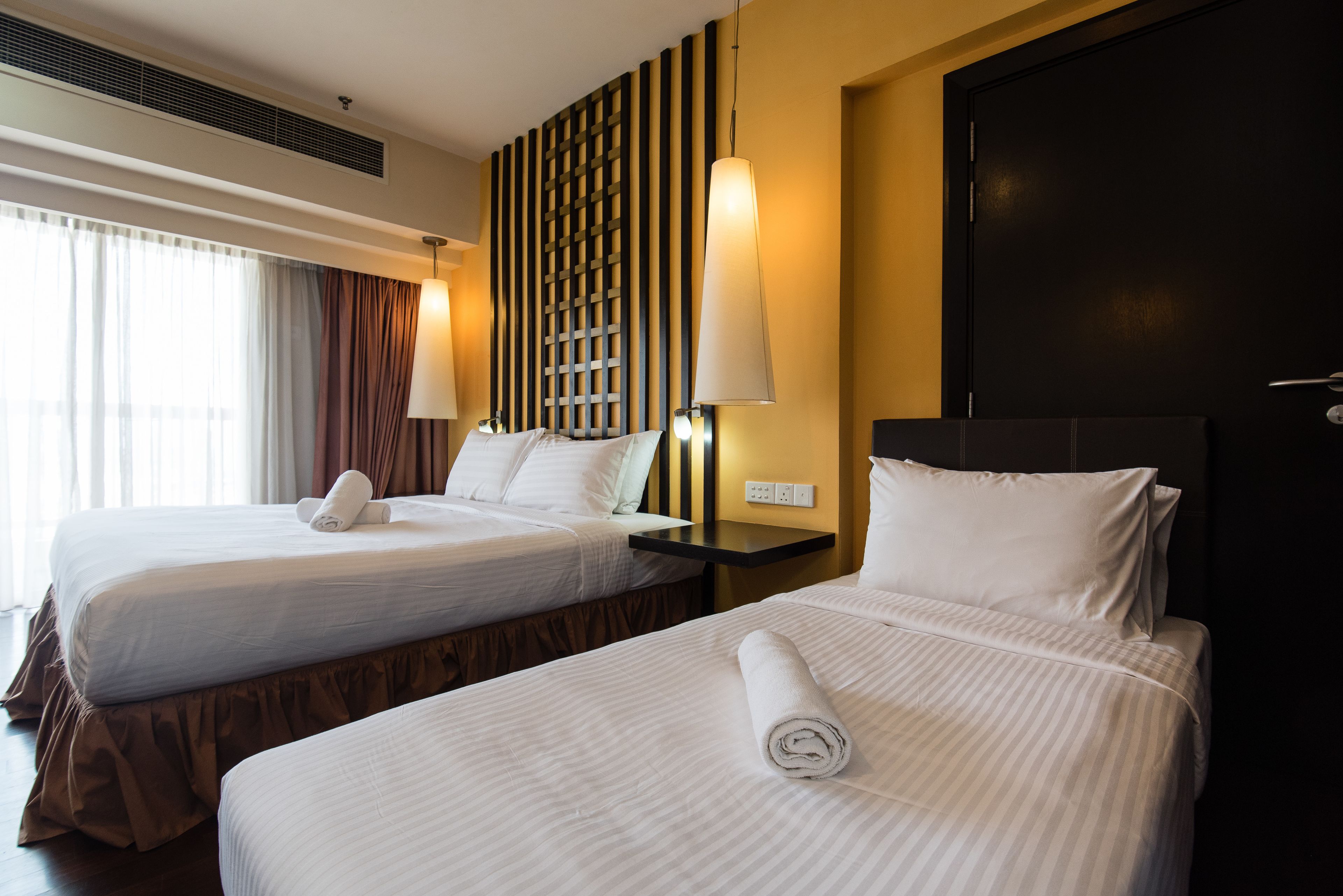 Dos camas de hotel contiguas con sábanas blancas, un cabecero de madera oscura y una lámpara fijada a la pared.