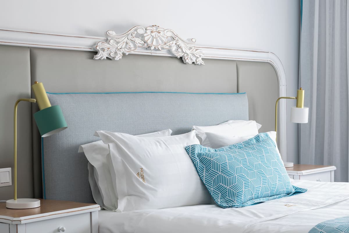 A neatly made bed with white bedding and a teal geometric pillow, flanked by two bedside tables with lamps, in a modern room with a decorative headboard.