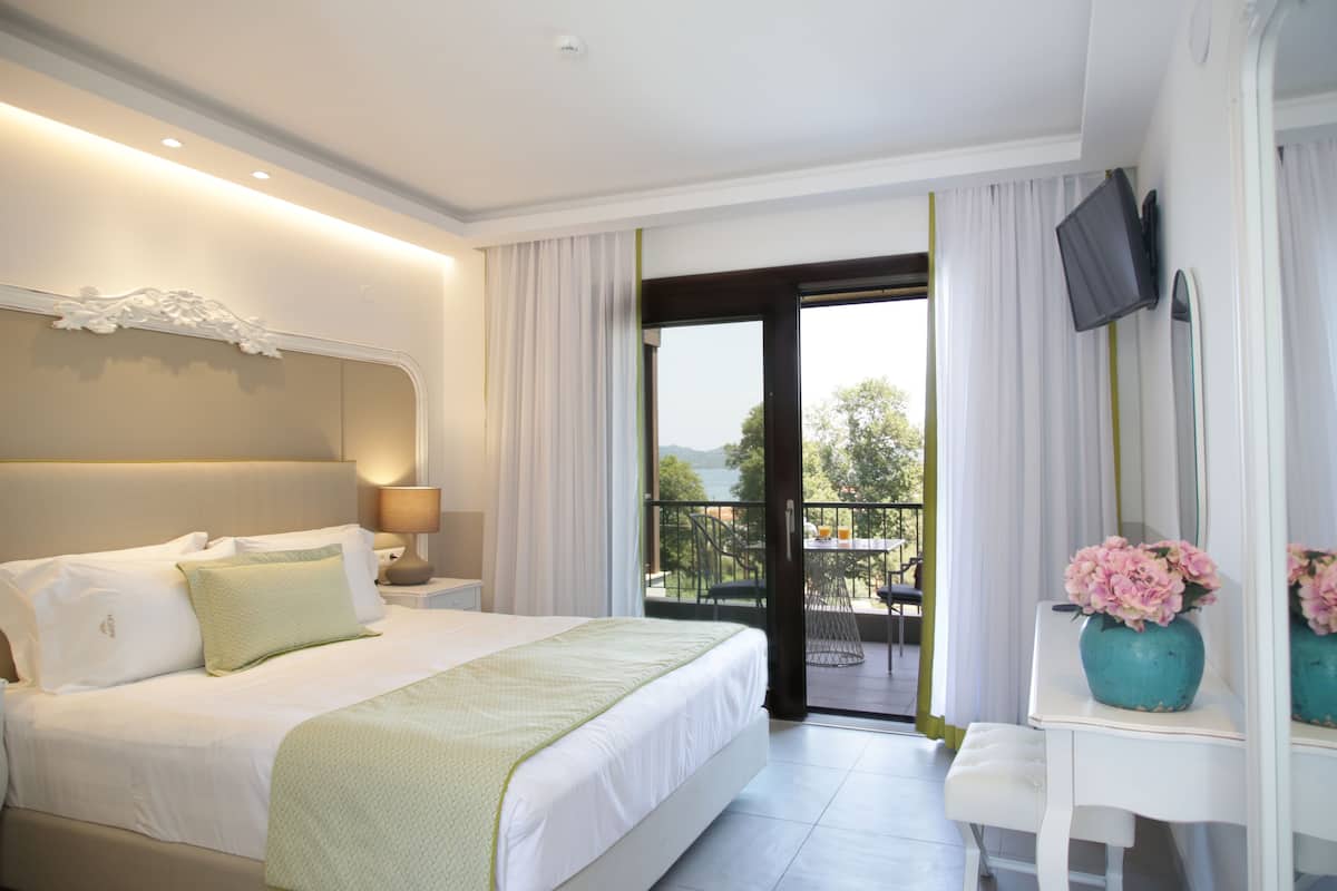 A hotel room with a large bed, a desk with a vase of flowers, a TV mounted on the wall, and a balcony with a view.