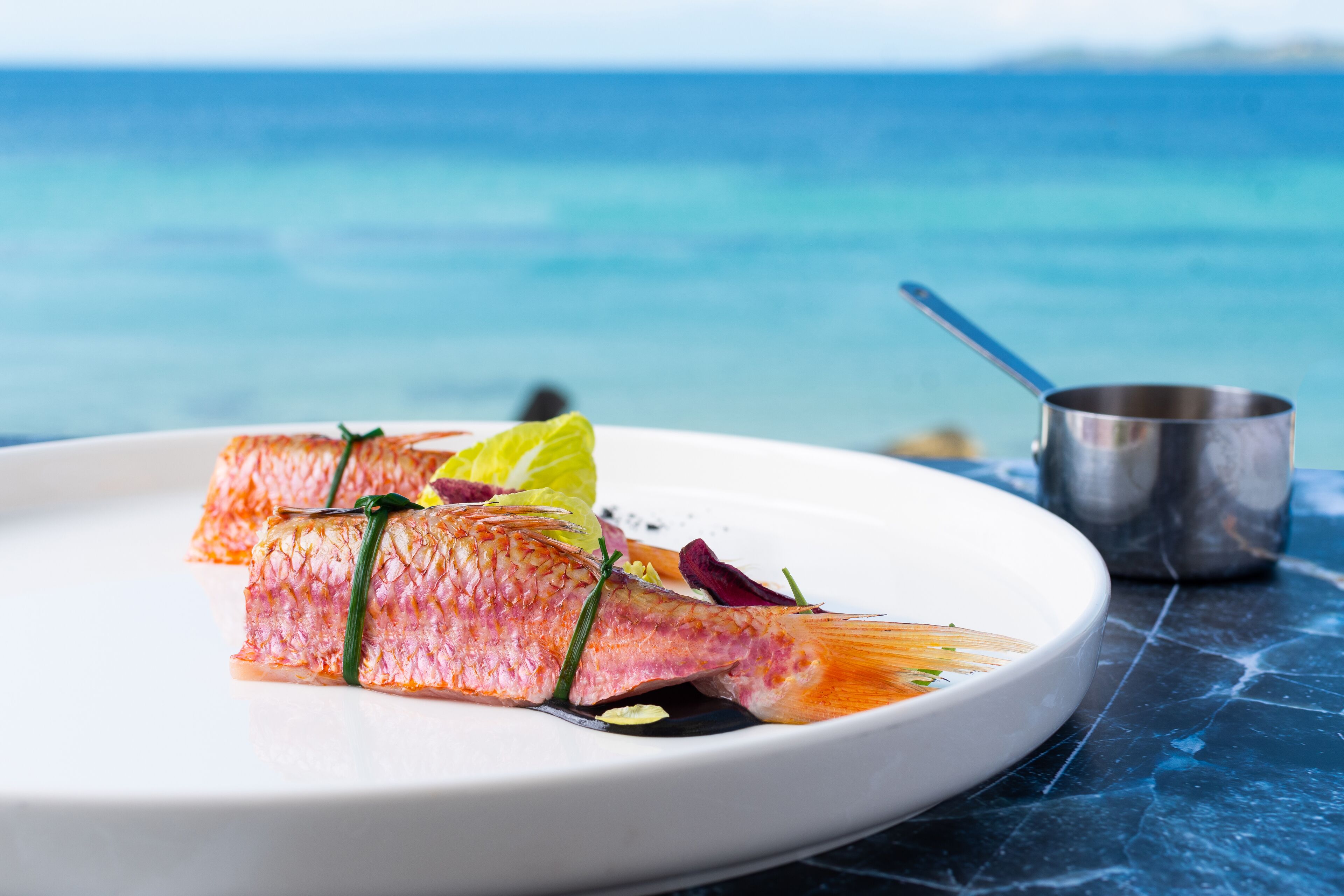Une assiette remplie de nourriture, avec un poisson comme élément central, un petit gobelet en métal et une surface en marbre bleu.