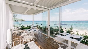 A covered outdoor dining area with tables and chairs, overlooking a beach and the sea. - Avaton Luxury Beach Resort - Relais & Chateaux (Aristotelis)