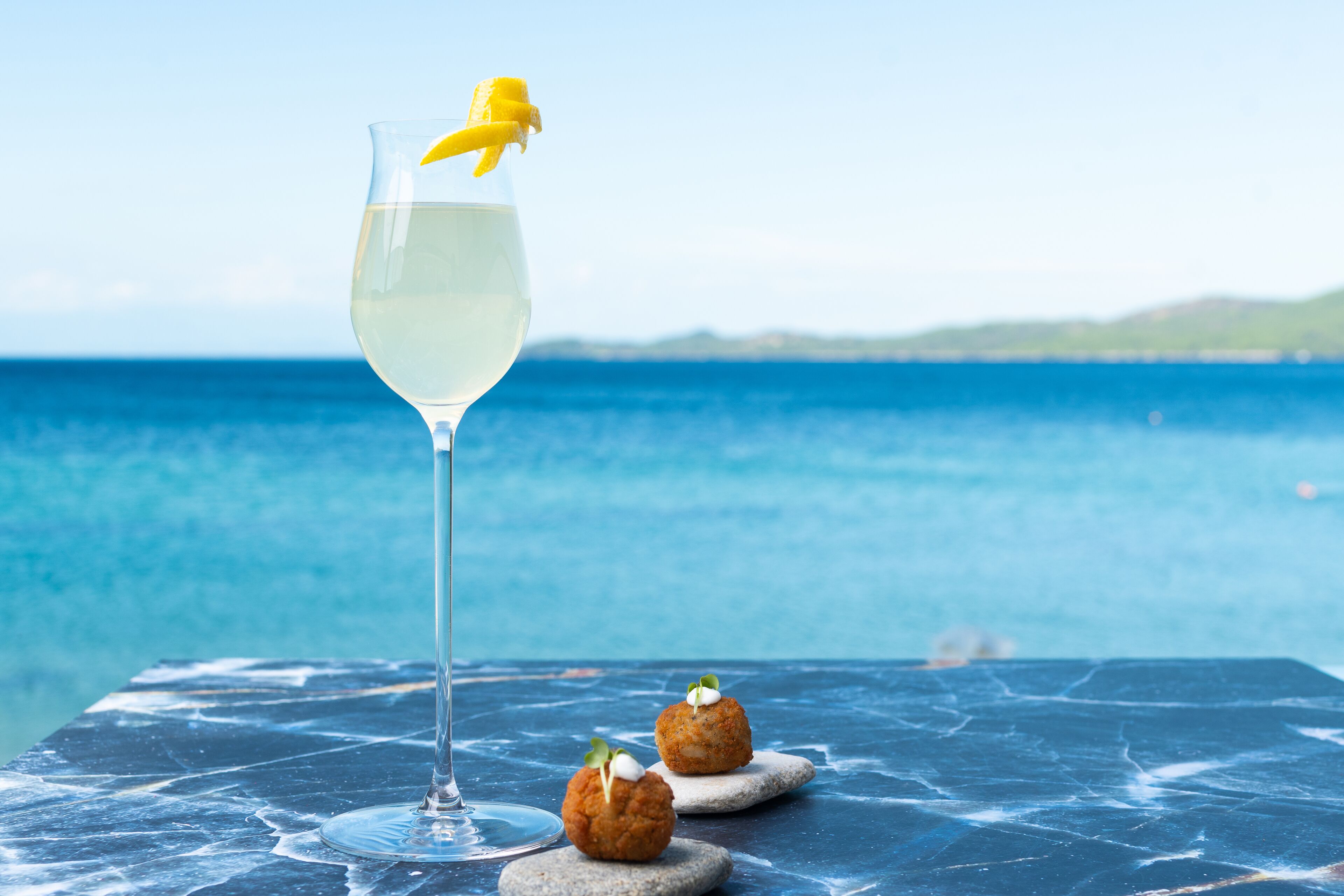 Une table sur laquelle se trouve un verre transparent contenant une boisson claire et de couleur pâle, deux petits desserts ronds recouverts d’une crème blanche, et une pelure de banane servant de décoration. L’arrière-plan représente une mer calme et une côte lointaine.