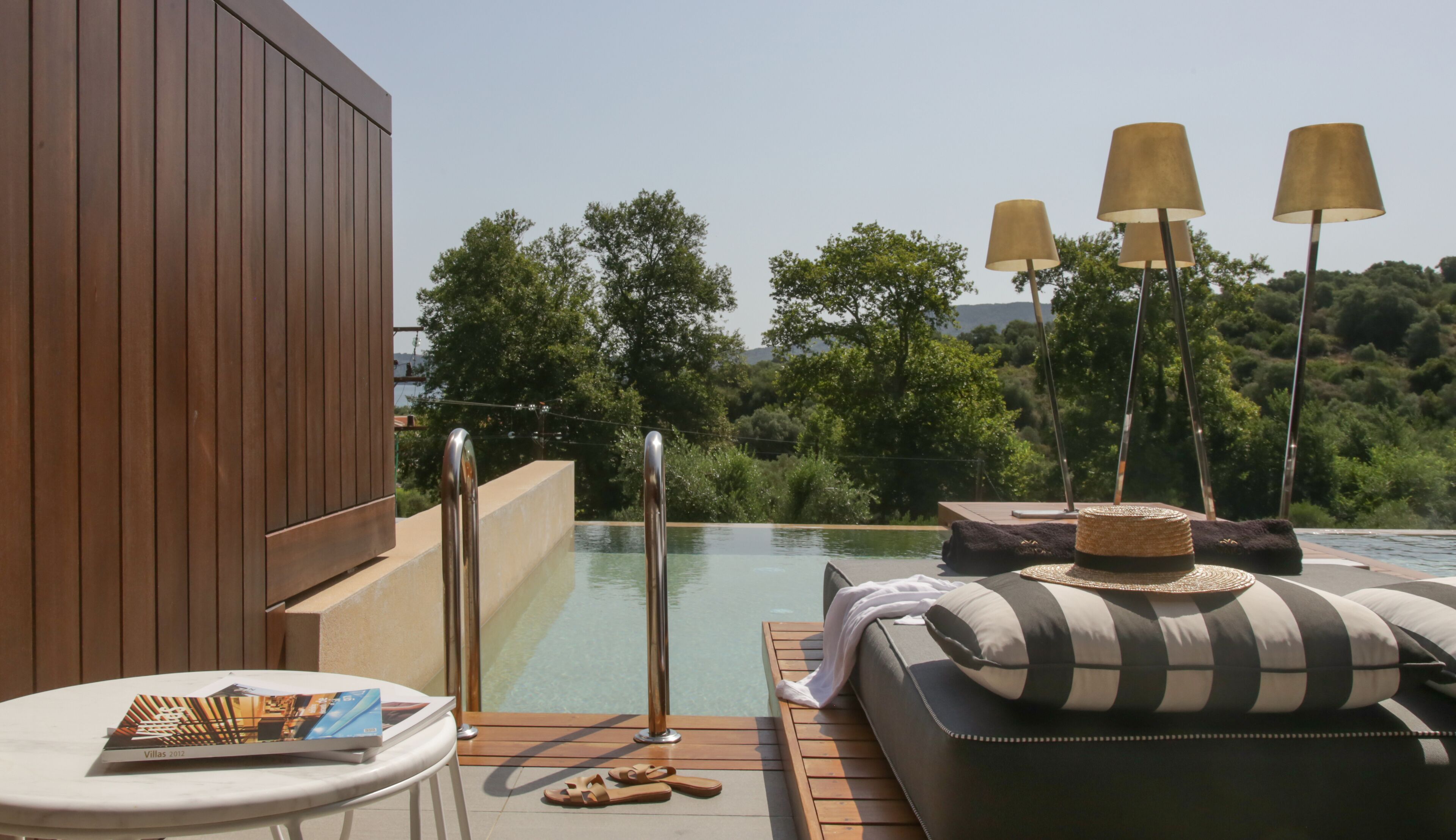 Zona de piscina exterior con terraza de madera, sillas de descanso y vista a un paisaje boscoso.