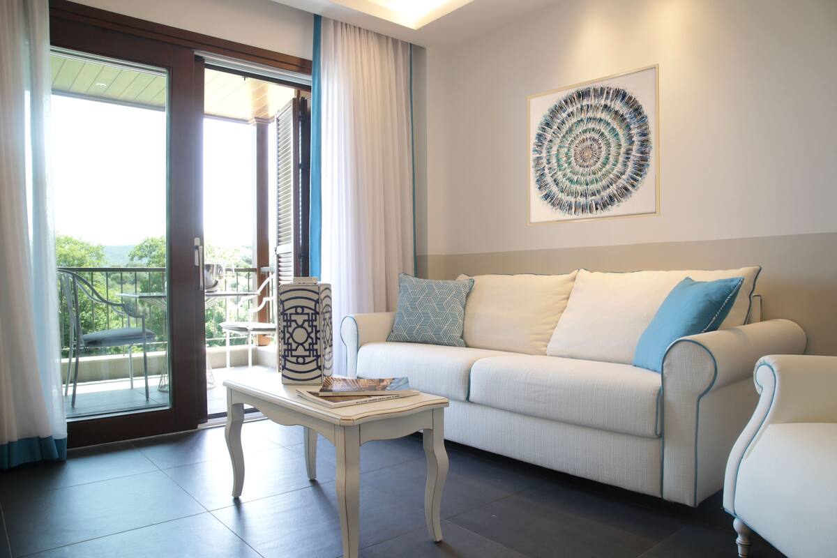 A modern living room with a white sofa, a coffee table, and a large window with a view of greenery.