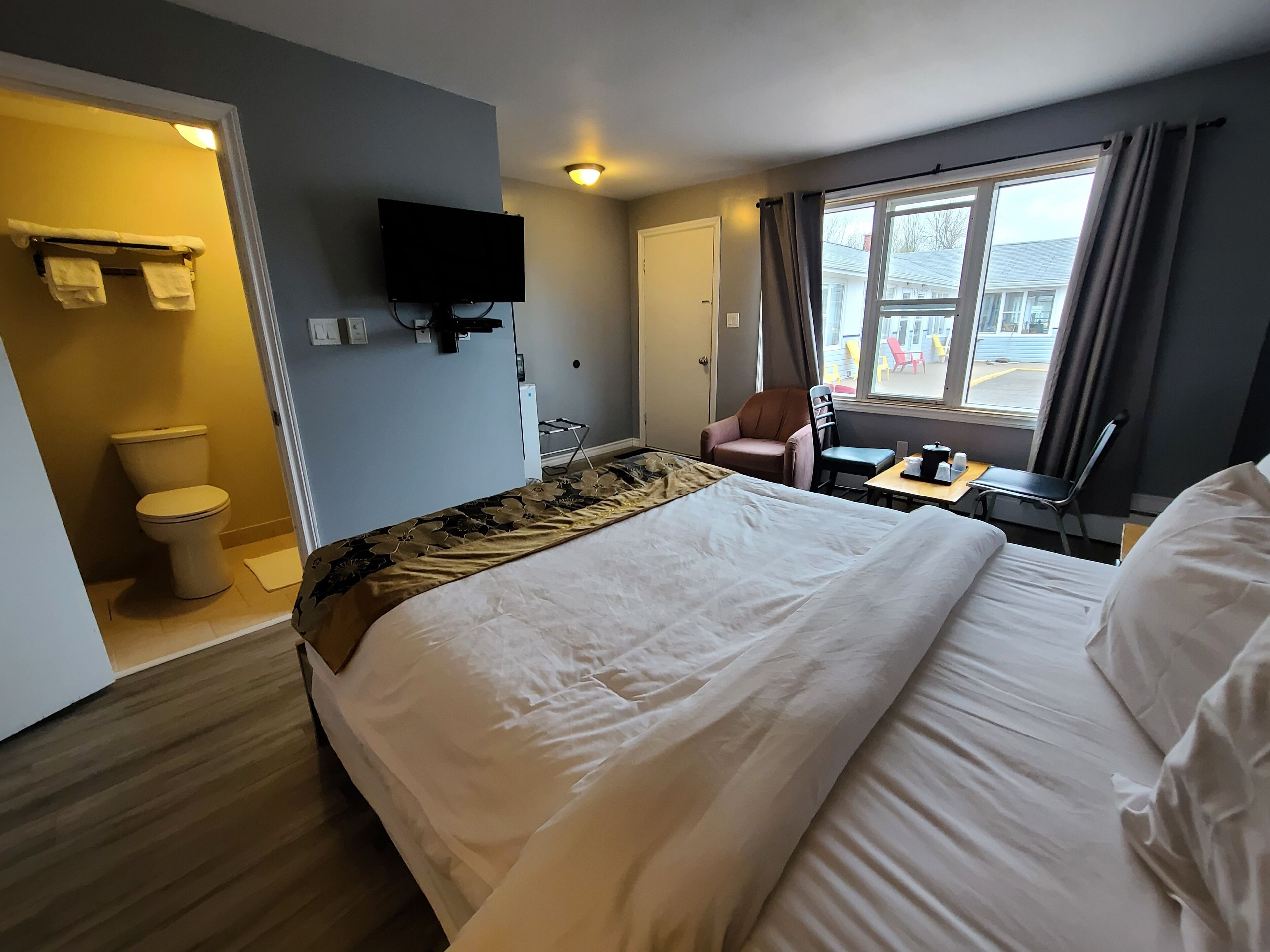 Habitación de hotel con una cama grande, un televisor fijado en la pared, una mesita con una silla y una ventana con cortinas.