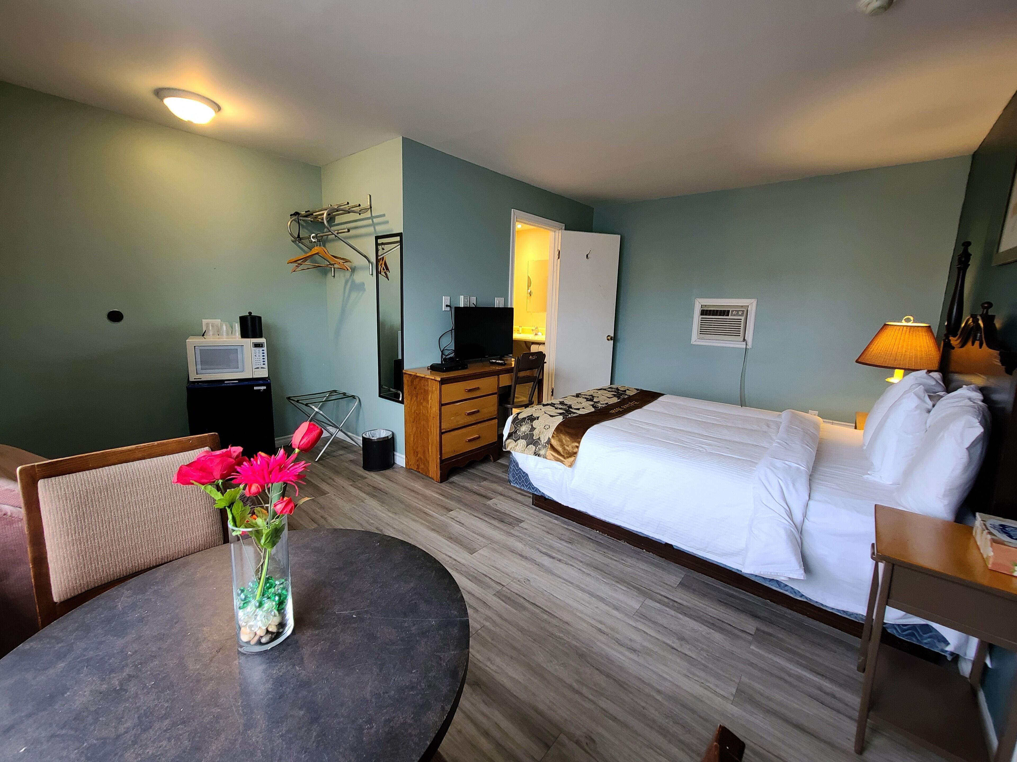 A hotel room with a bed, a desk, a chair, a small table with a vase of flowers, a television, and a lamp.