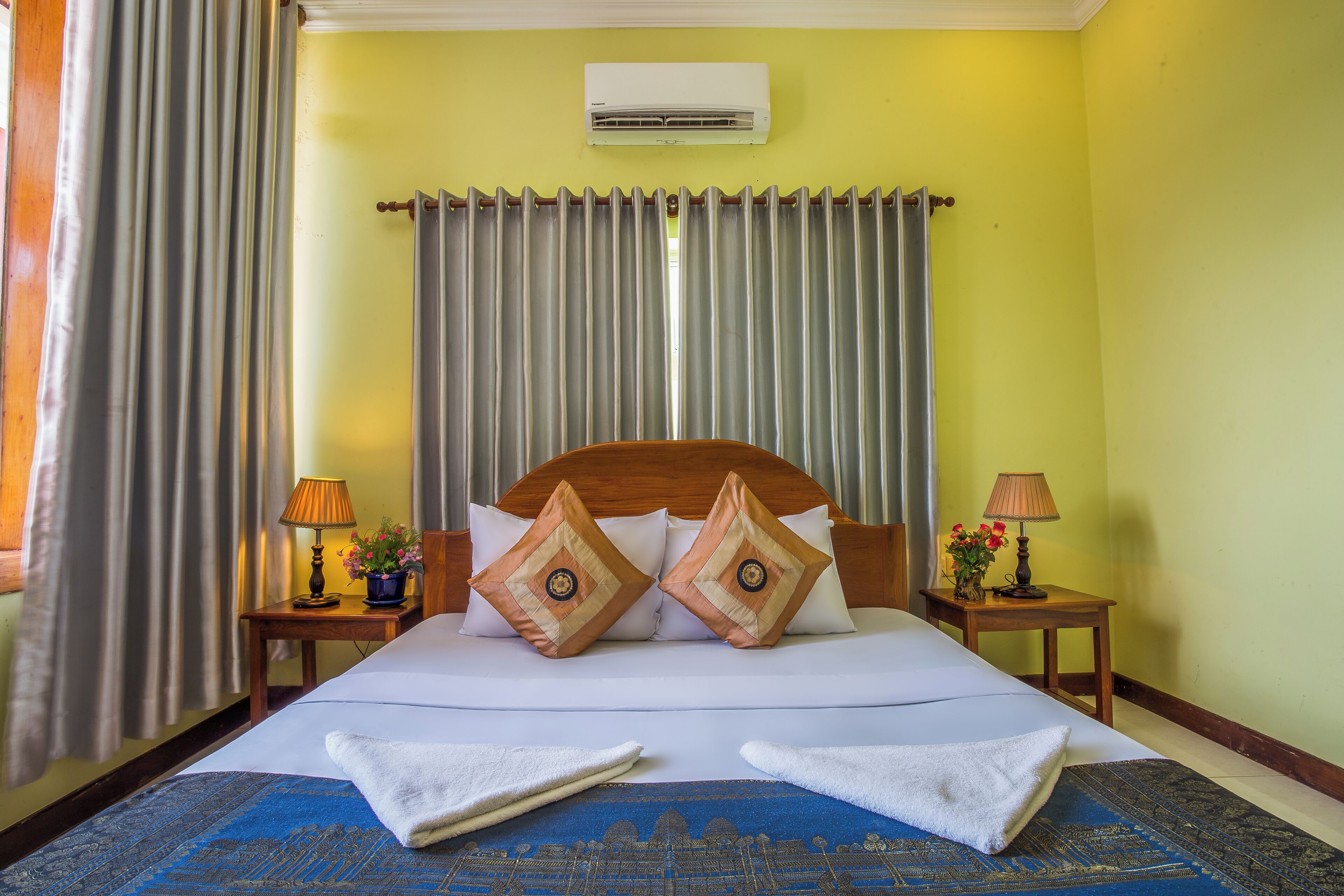 A hotel room with a bed, two nightstands, a window with curtains, an air conditioning unit, and a blue patterned rug.