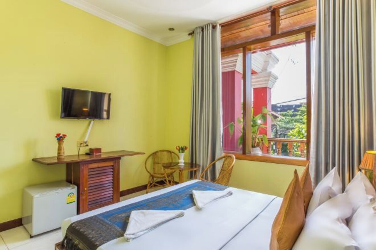 A hotel room with a bed, a TV mounted on the wall, a small desk with a chair, and a window with curtains.