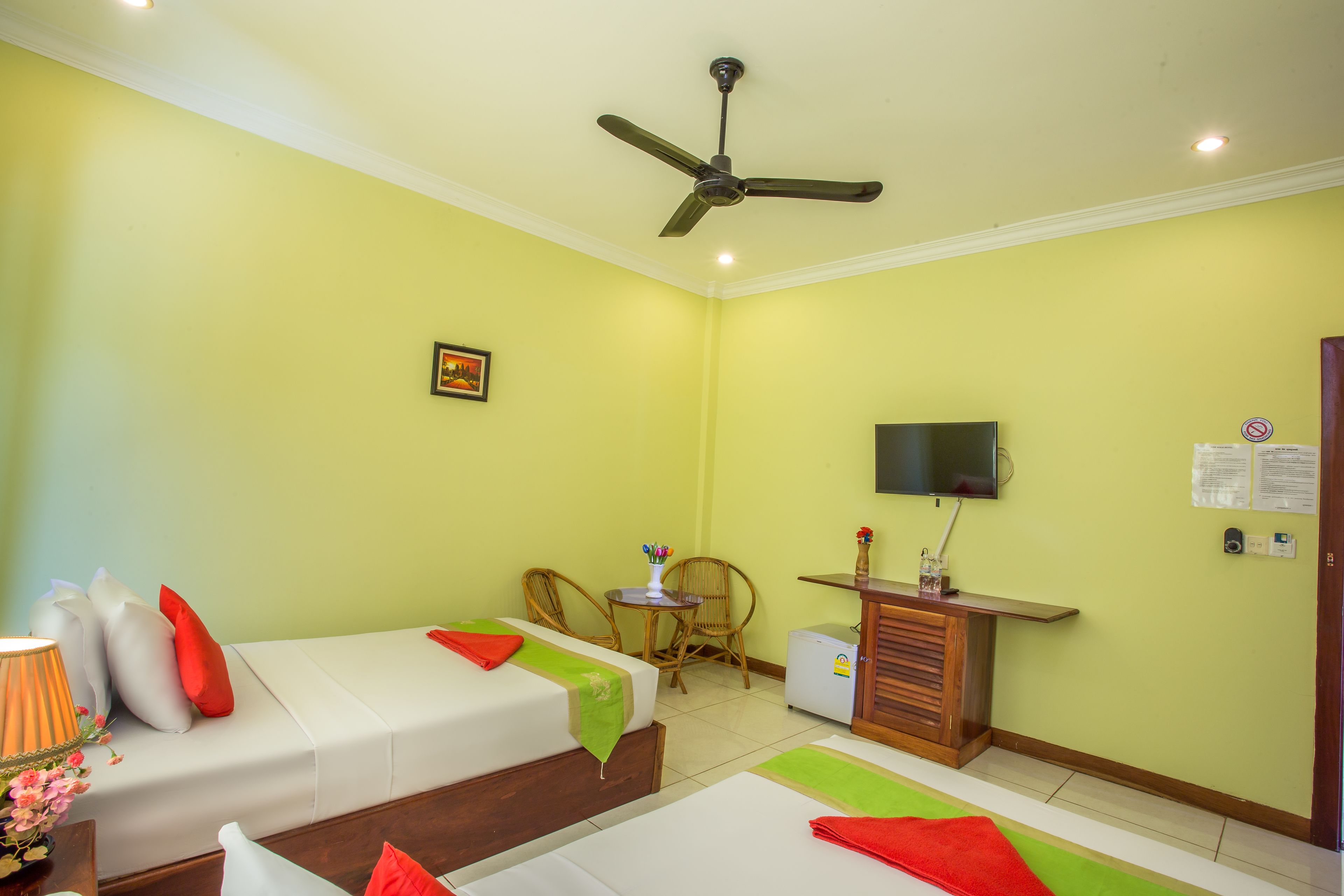 Habitación de hotel con dos camas, ventilador de techo, televisor, una mesita con un florero y un armario.