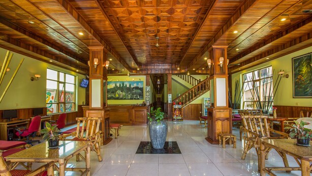 A lobby with a wooden ceiling, a large vase with plants, and a staircase. - Side Walk Never Die (Siem Reap)