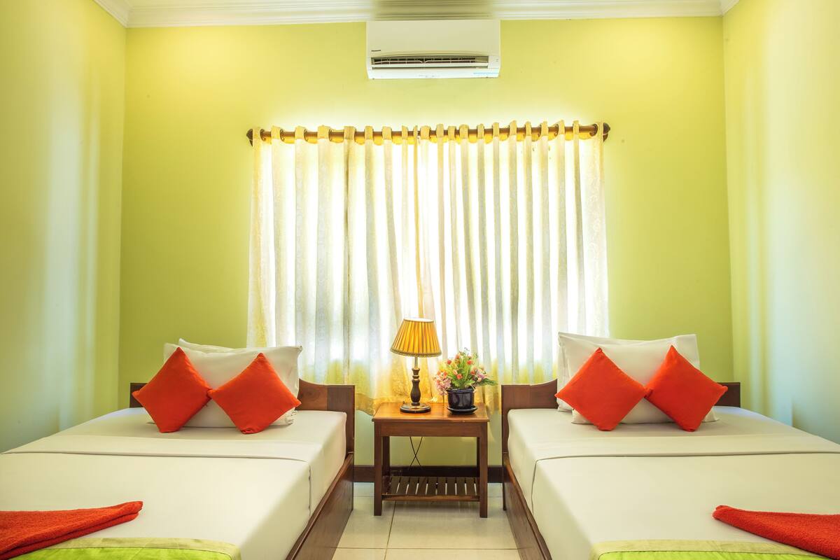 A hotel room with two beds, a small table with a lamp and a flower pot, and a window with curtains.