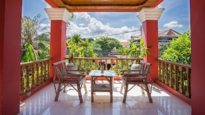 A covered balcony with two wooden chairs and a table, surrounded by red walls and a view of greenery and buildings. - Side Walk Never Die (Siem Reap)