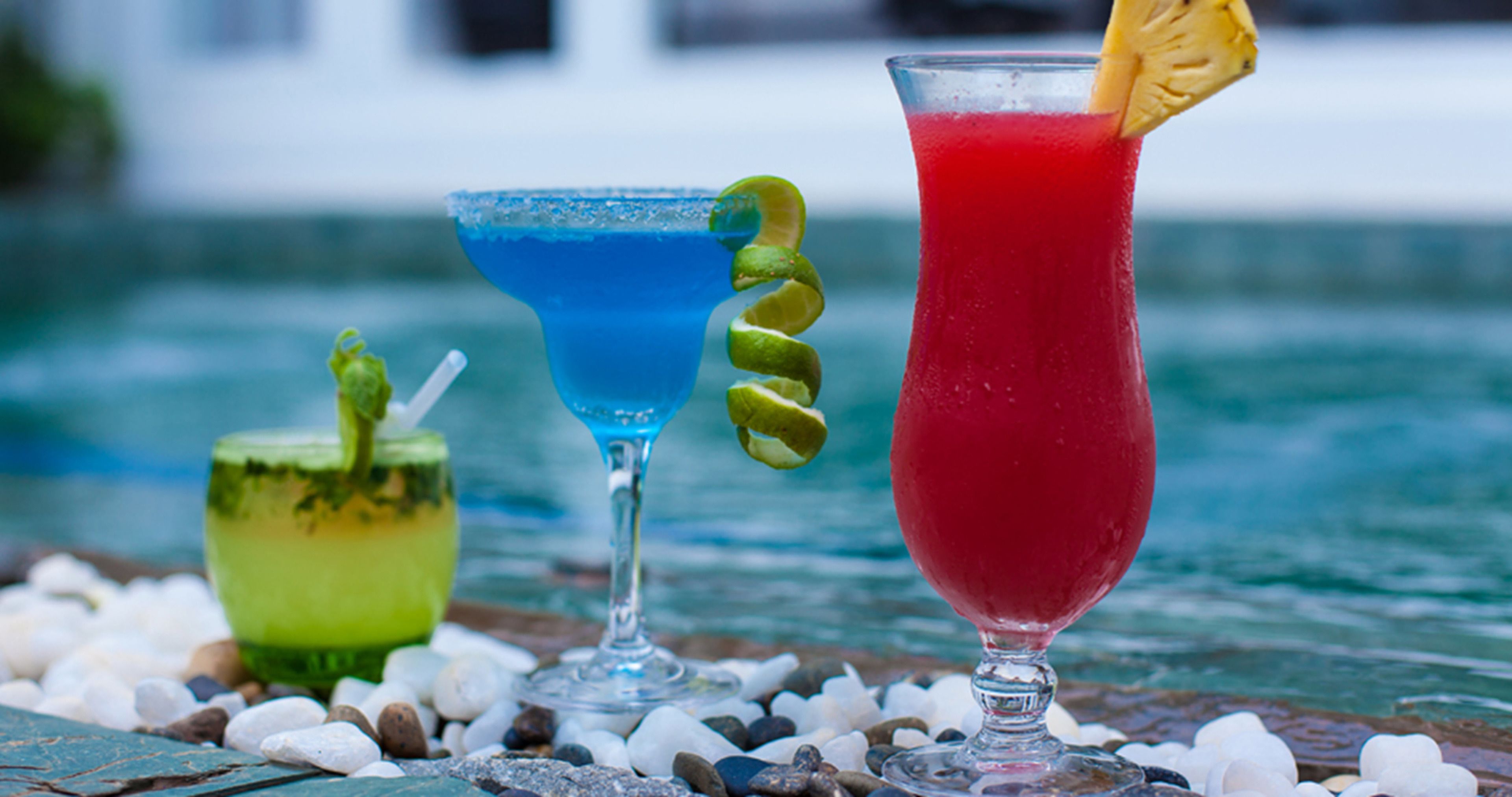 Three cocktails on a table with pebbles, near a body of water.