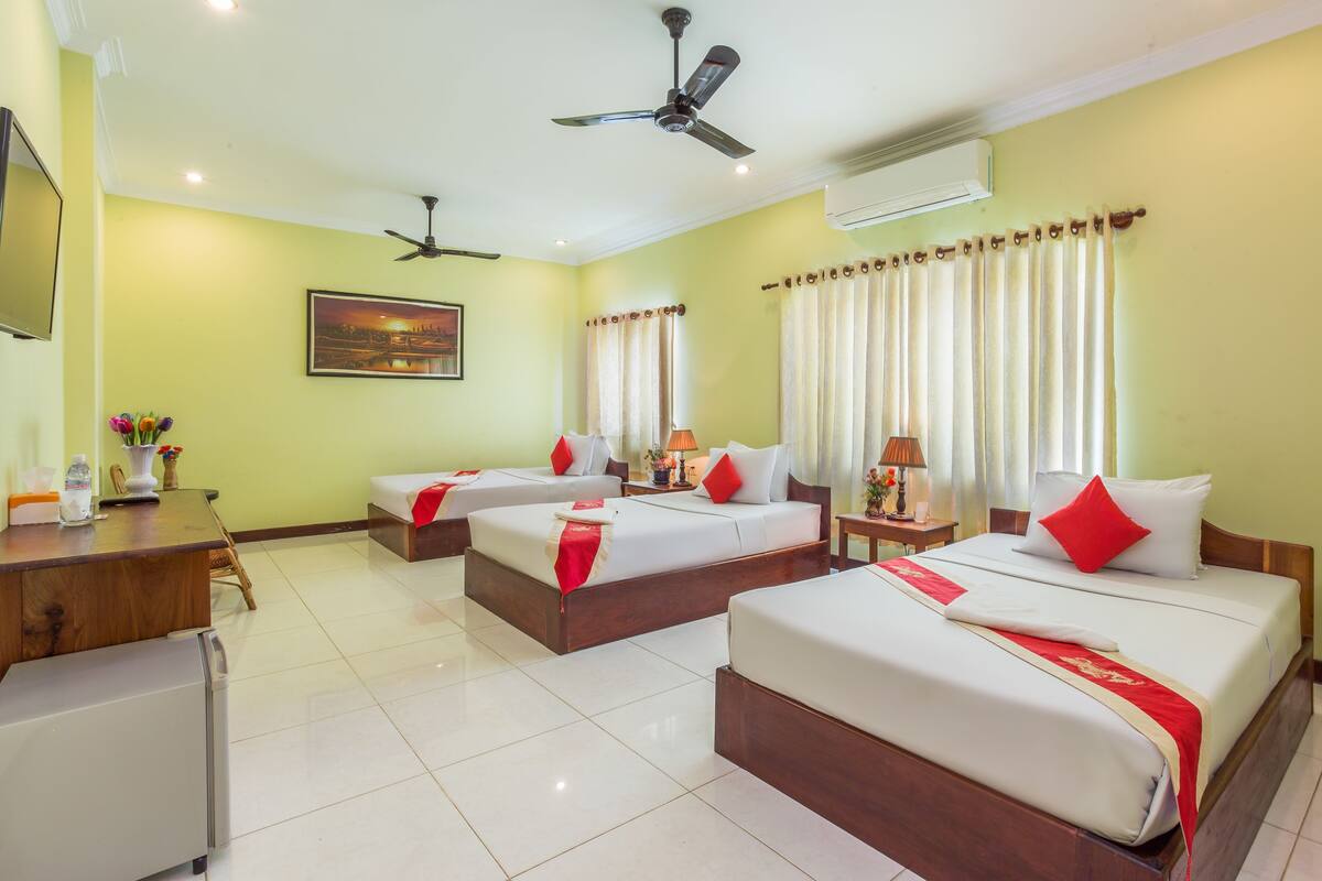 A hotel room with three beds, a desk, a refrigerator, and a ceiling fan.