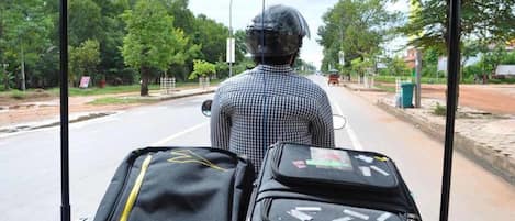 Una persona sentada en un vehículo con equipaje, mirando una carretera bordeada de árboles bajo un cielo despejado.