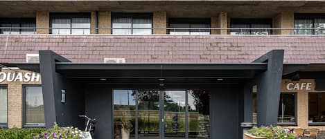 Entrada de un edificio con puerta de vidrio, dos grandes macetas de madera con flores, y letreros que dicen 'QUASH' y 'CAFE'.
