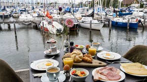 Une table de petit-déjeuner avec des assiettes d’œufs, des toasts, des croissants et un panier de fruits, le tout disposé devant un paysage de port de plaisance.