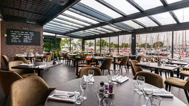 Un espace de restauration intérieur avec des tables dressées pour les clients, un tableau affichant le menu et une vue sur les voiliers à travers de grandes fenêtres.