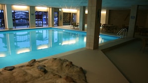 Une piscine intérieure à l’eau claire, entourée de chaises longues et de tables, et une grande formation rocheuse en premier plan.