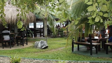 An outdoor seating area with thatched-roof tables and chairs, surrounded by lush greenery and palm trees.