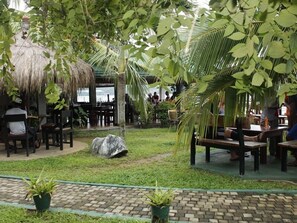An outdoor seating area with thatched-roof tables and chairs, surrounded by lush greenery and palm trees.
