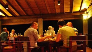 Een groep mensen dineert aan een houten tafel in een kamer met weinig licht, hoge plafonds en zichtbare balken.