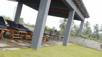 A covered outdoor area with lounge chairs and a table, surrounded by palm trees and a concrete wall.