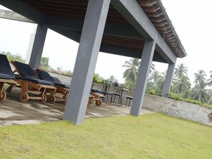 A covered outdoor area with lounge chairs and a table, surrounded by palm trees and a concrete wall.