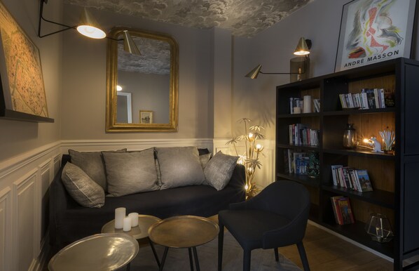 A cozy living room with a dark sofa, a small round table, and a bookshelf filled with books. - Hôtel Jeanne d'Arc Le Marais (Paris)