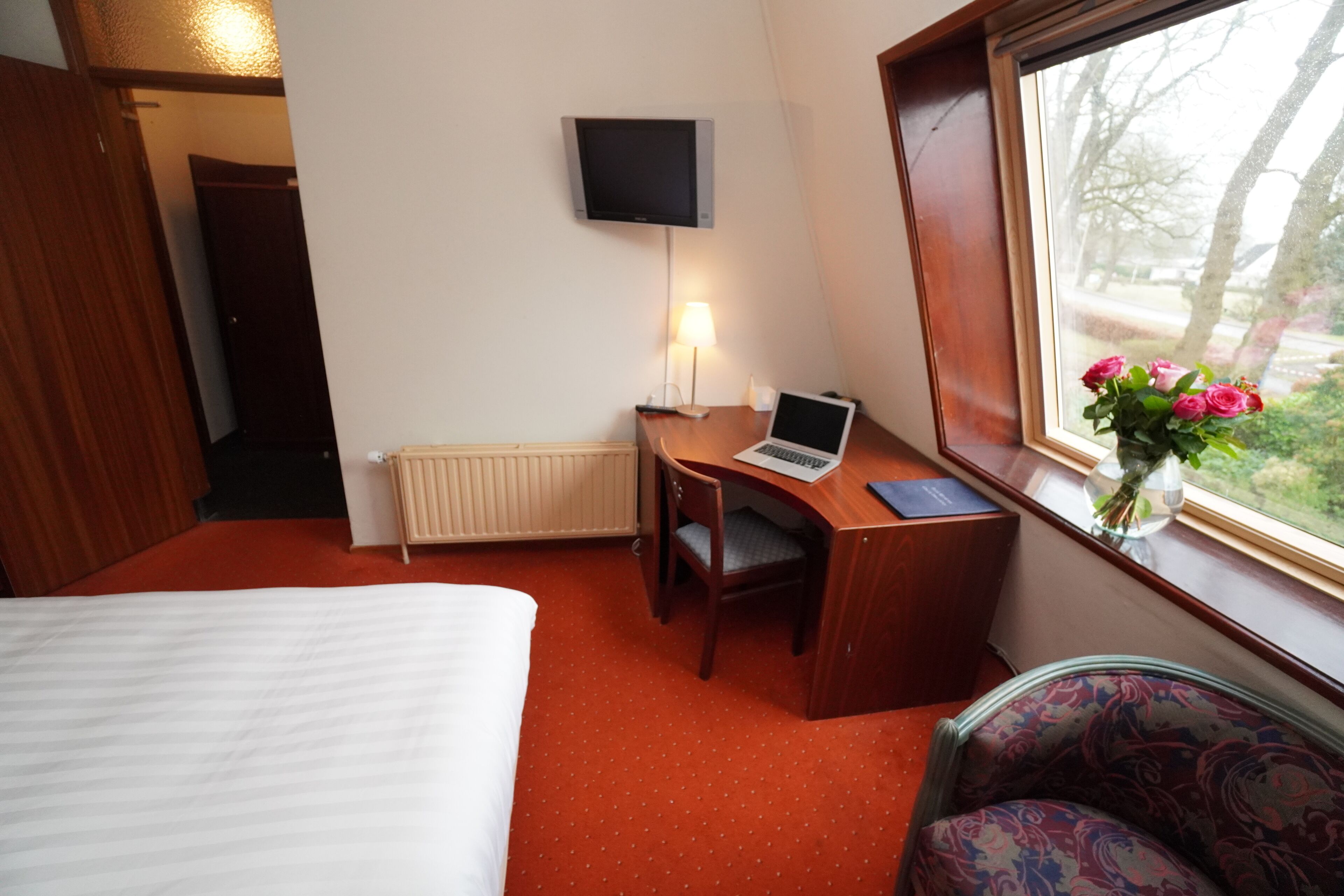 A hotel room with a bed, a desk with a laptop, a chair, a TV on the wall, and a window with a view of trees.