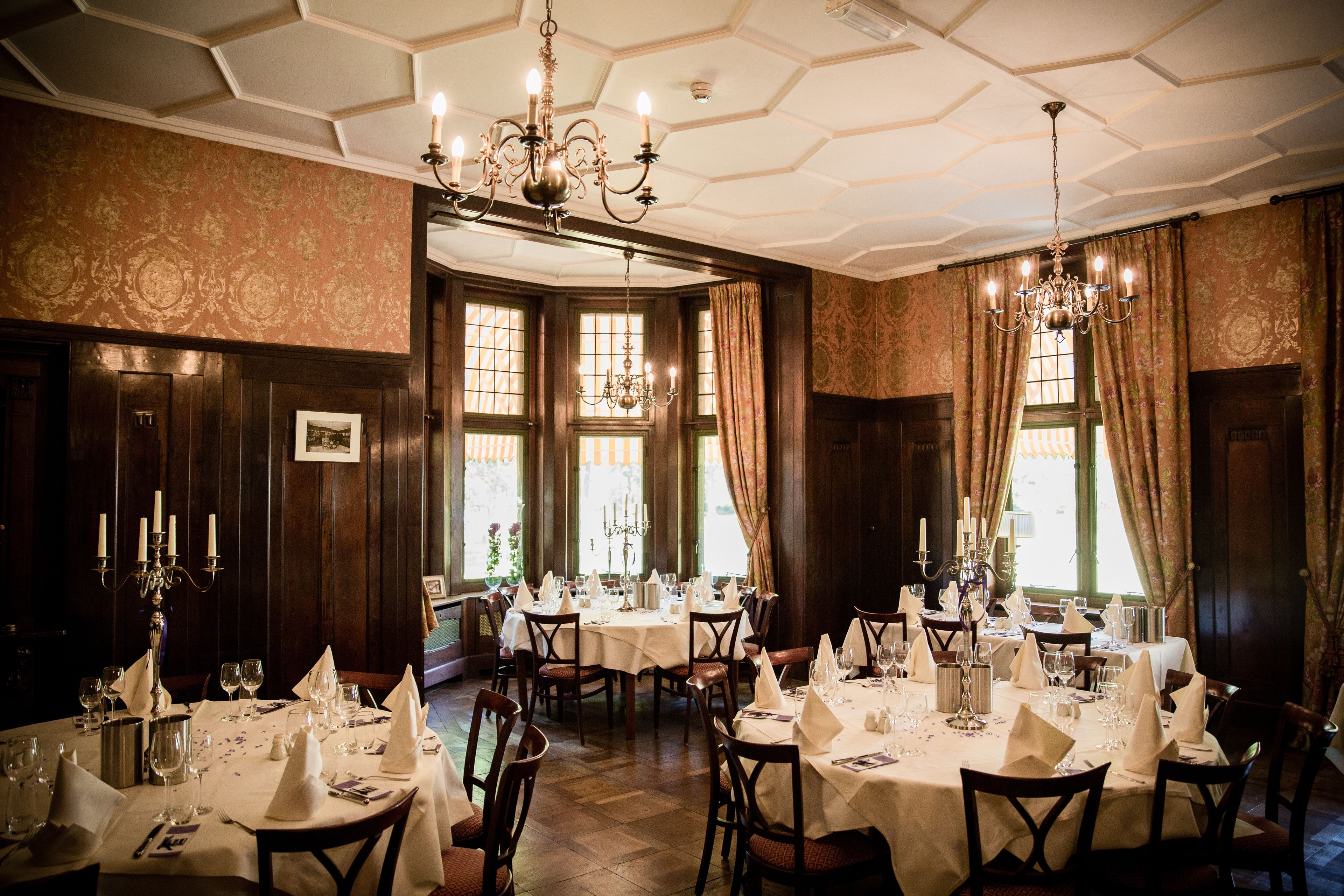 Un comedor elegante con varias mesas dispuestas para una comida, ventanas amplias con cortinas y una araña.
