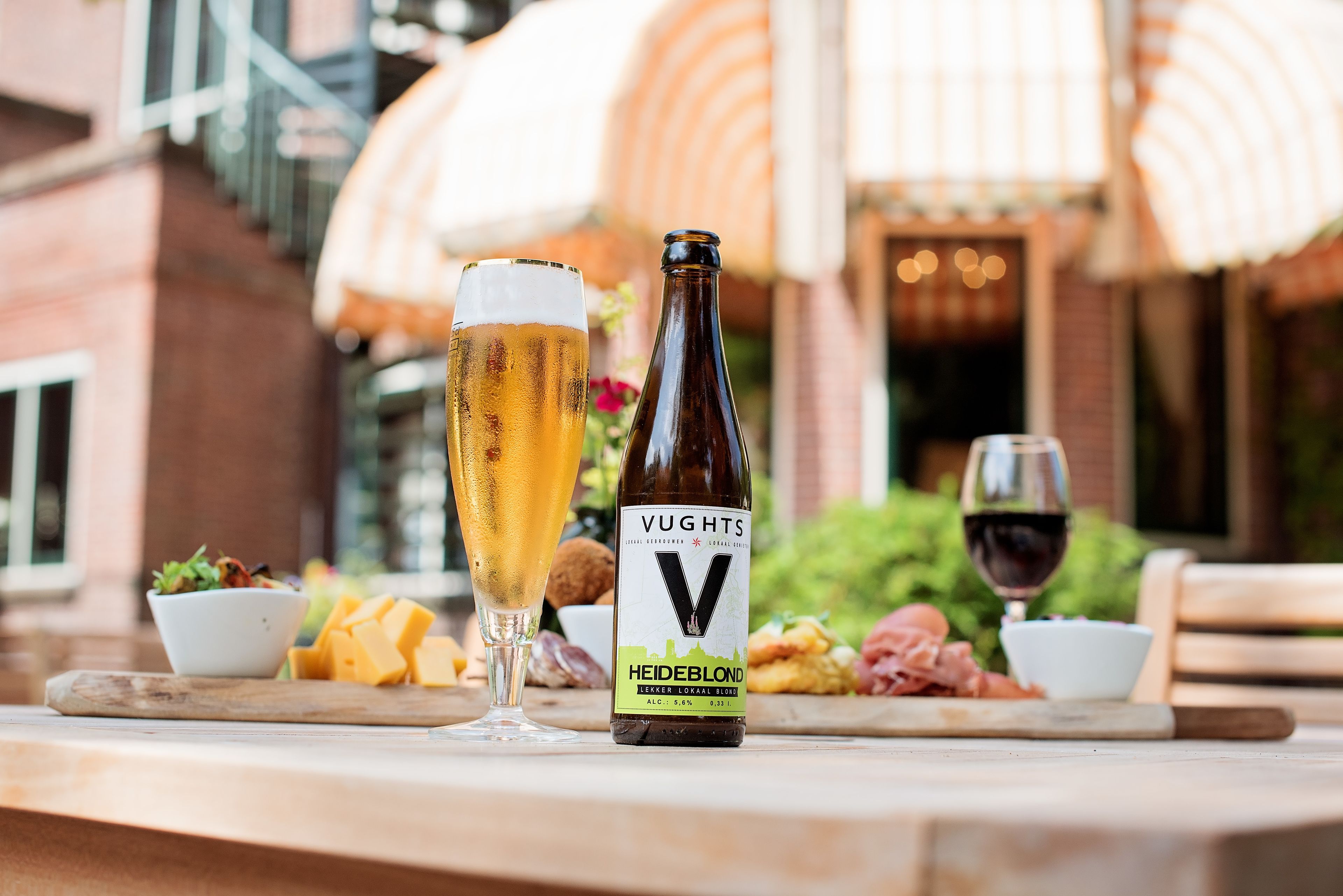 Una botella de cerveza Vughts Heiderblond, un vaso de cerveza y un vaso de vino tinto sobre una mesa con comida.