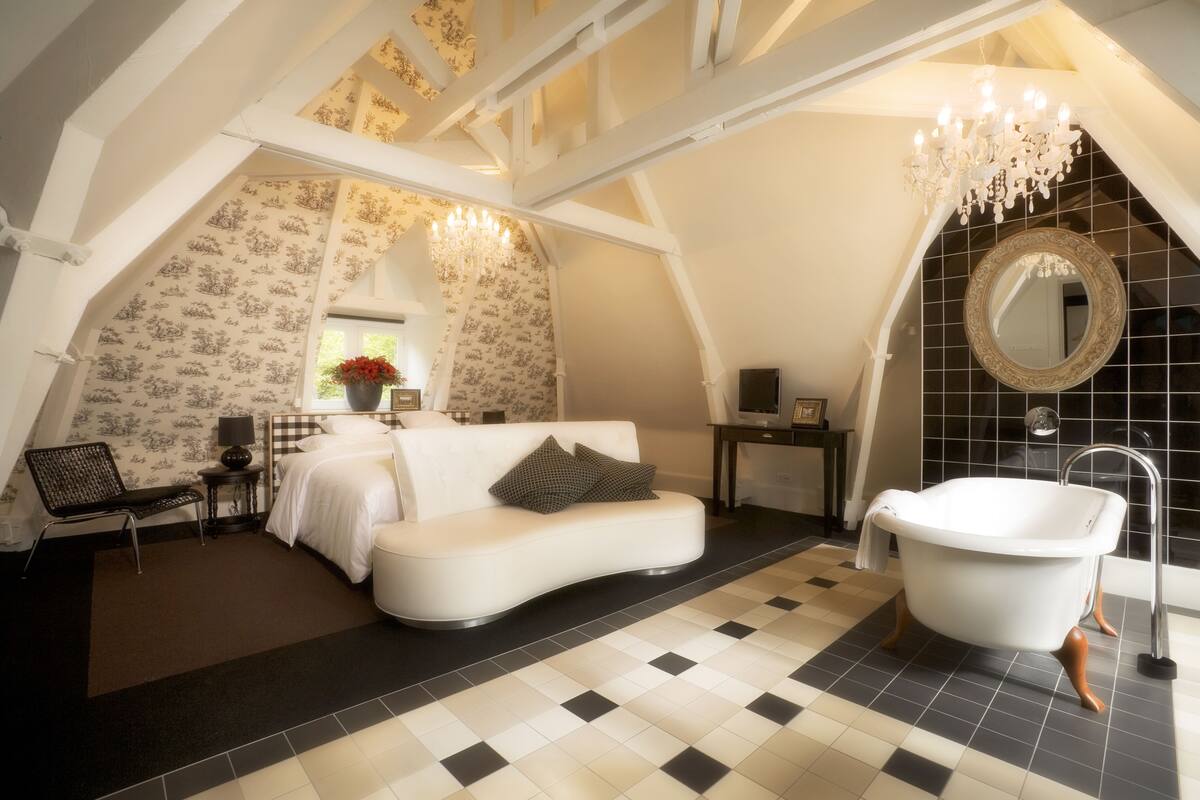 A bedroom with a white sofa, a white bathtub, a chandelier, and patterned wallpaper.