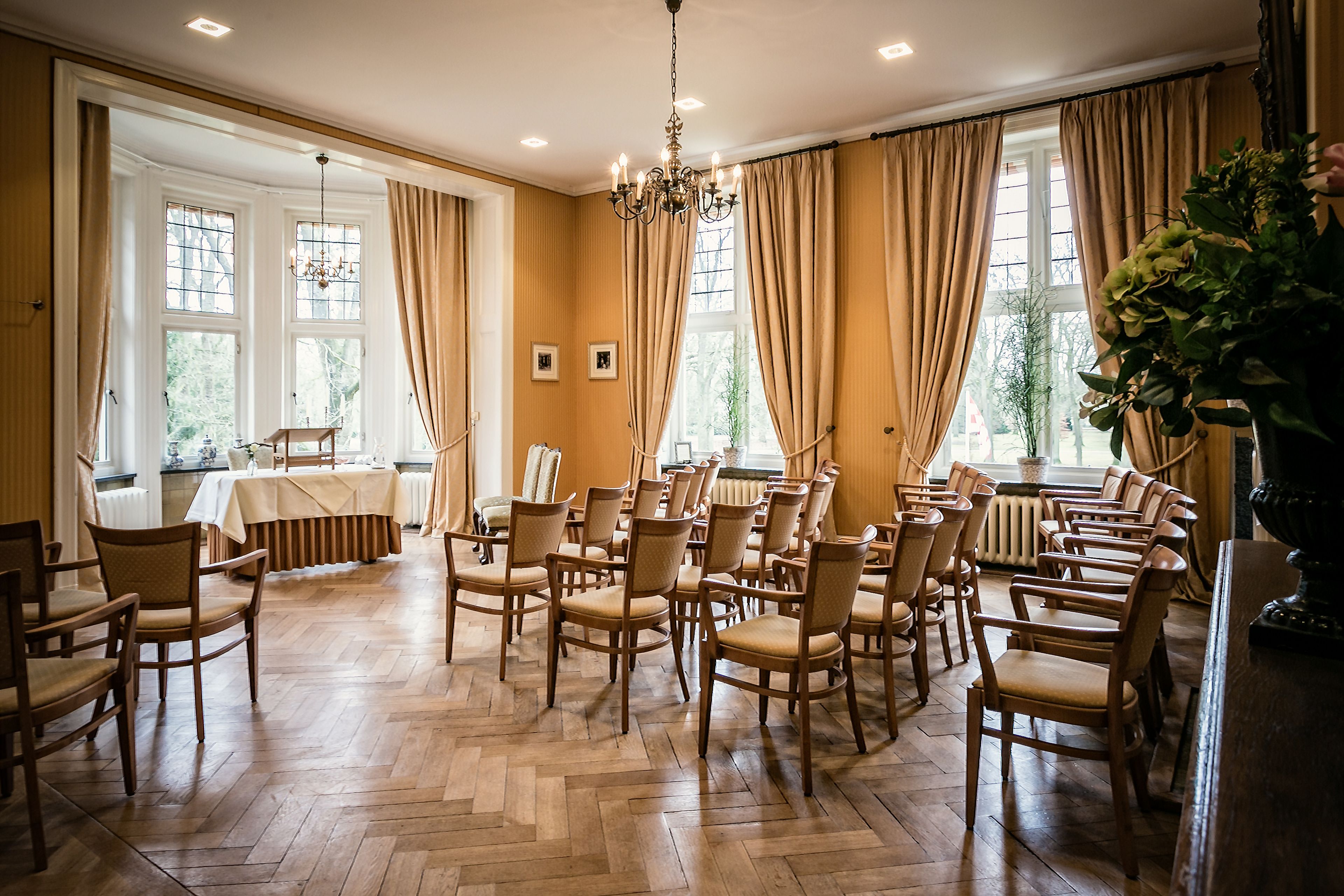 A room with wooden chairs arranged in rows, large windows with curtains, and a chandelier.