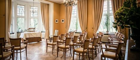 Una sala con sillas de madera dispuestas en filas, ventanas amplias con cortinas y un candelabro.