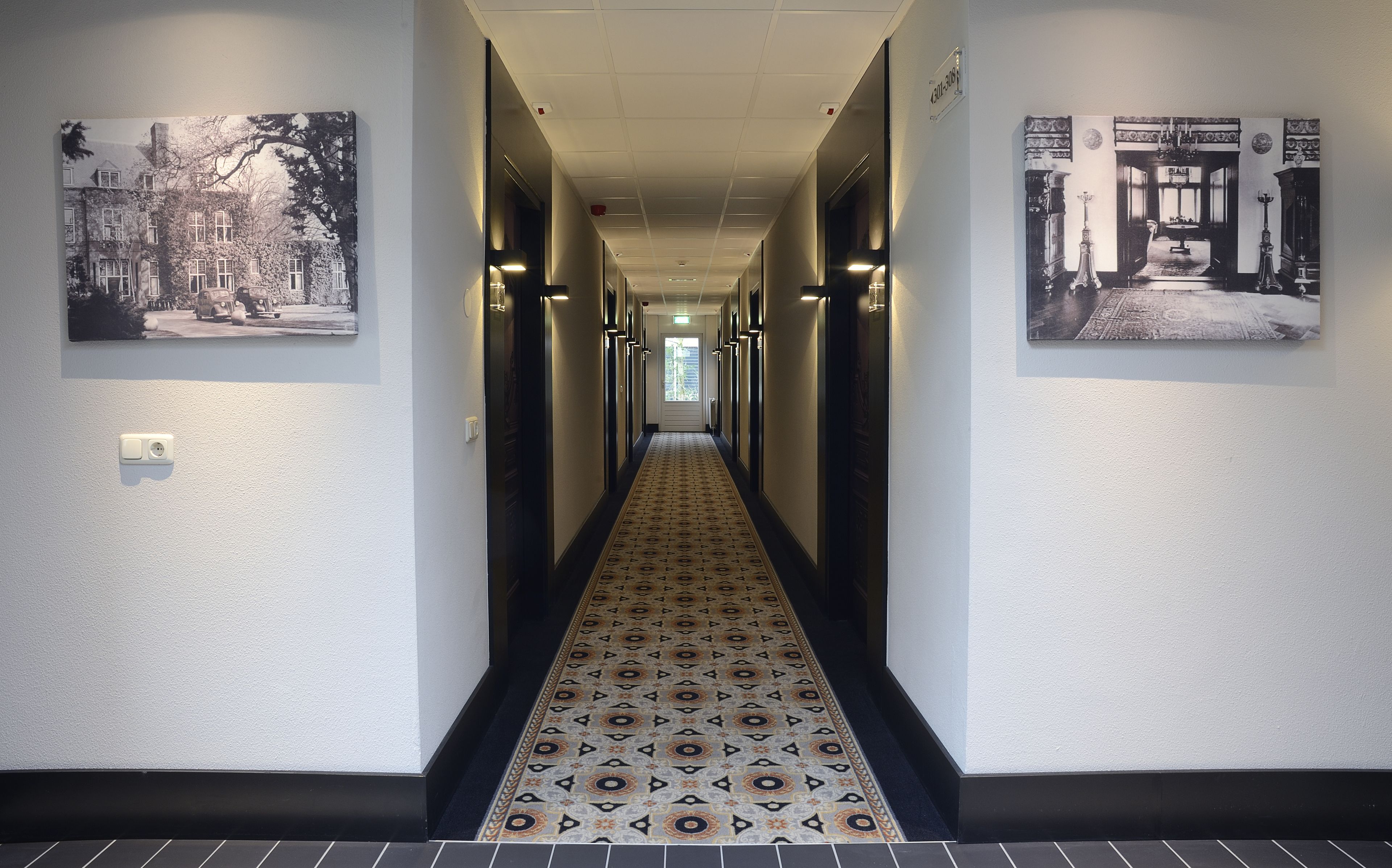 En hall med ett mönstrat golv, dörrar med svarta ramar och vägglampor med svarta ramar.
