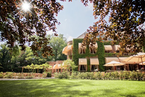 A building with a brick exterior and green ivy, surrounded by a well-maintained lawn and outdoor seating with umbrellas. - Landgoed Huize Bergen (Vught)