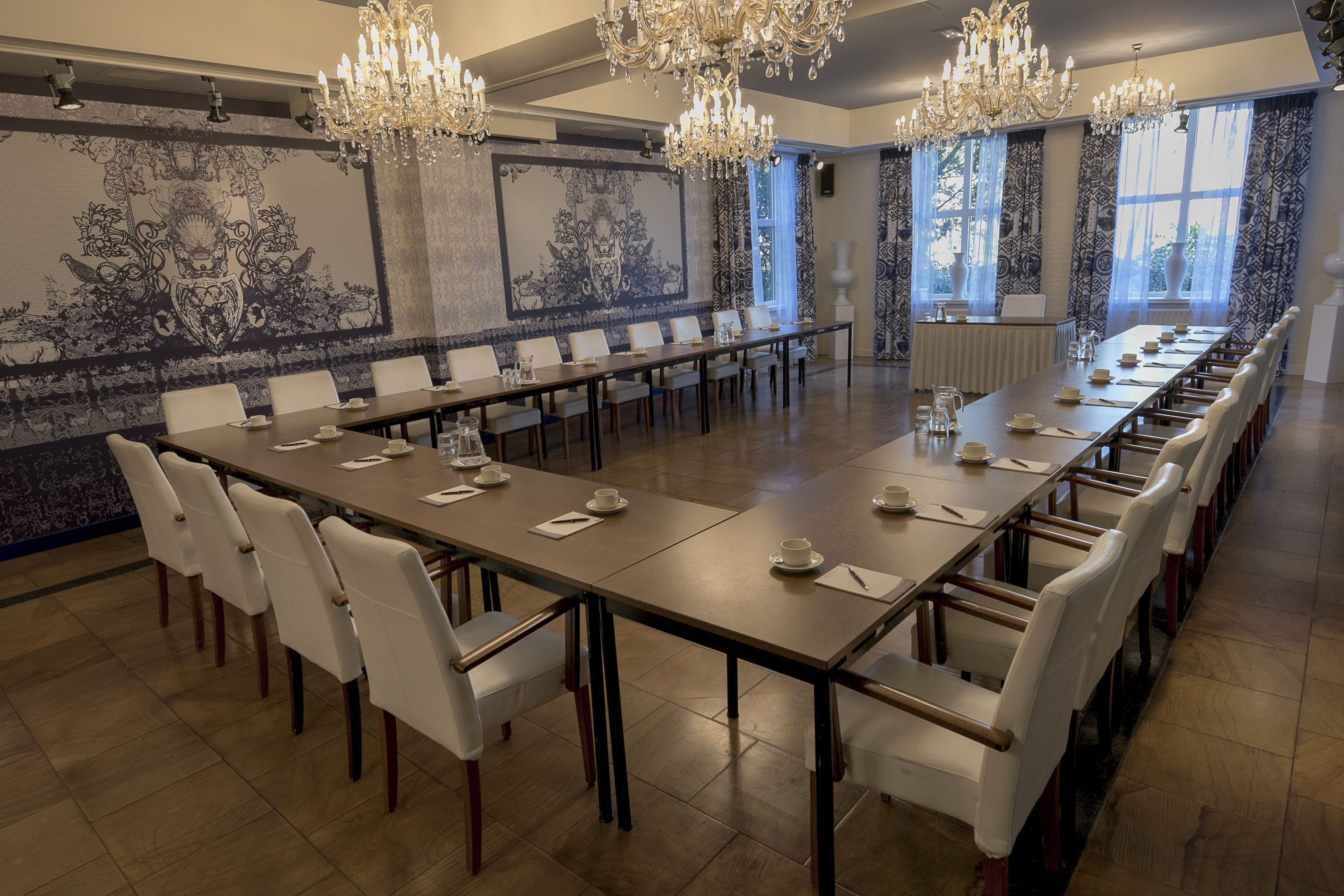 Un salón de conferencias con una larga mesa, sillas blancas, candelabros y decoraciones murales estampadas.