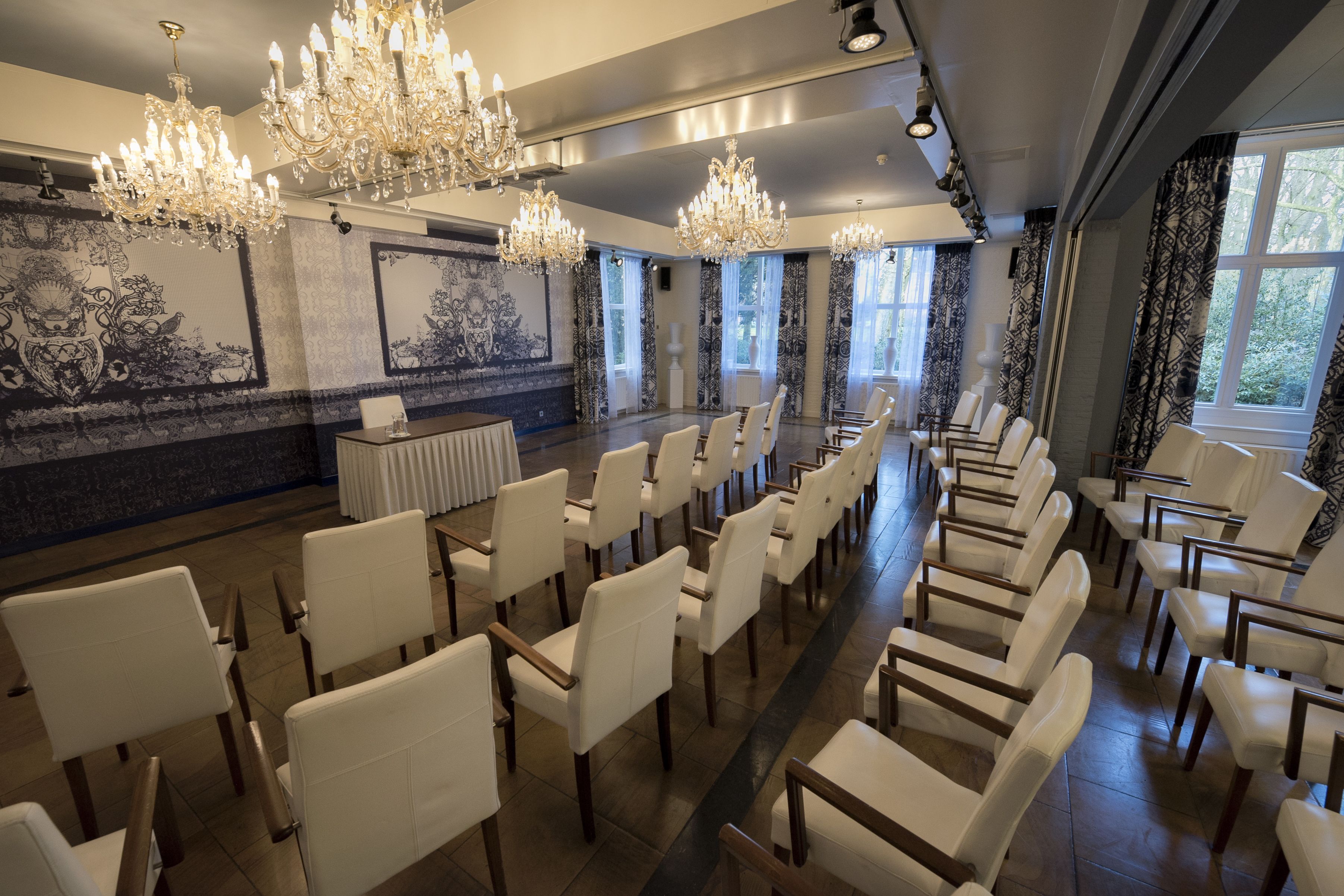 A conference room with rows of white chairs, chandeliers, and large windows with curtains.